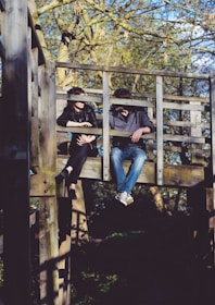 Two individuals are sitting on an elevated wooden platform structure in what appears to be a park or wooded area. They are having a conversation, with one person leaning on the railing and the other sitting casually with legs hanging off the platform. The background shows trees with sparse leaves, indicating it might be early spring or autumn.