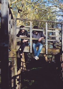 Two individuals are sitting on an elevated wooden platform structure in what appears to be a park or wooded area. They are having a conversation, with one person leaning on the railing and the other sitting casually with legs hanging off the platform. The background shows trees with sparse leaves, indicating it might be early spring or autumn.