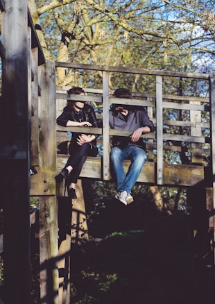 Two individuals are sitting on an elevated wooden platform structure in what appears to be a park or wooded area. They are having a conversation, with one person leaning on the railing and the other sitting casually with legs hanging off the platform. The background shows trees with sparse leaves, indicating it might be early spring or autumn.