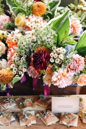 A vibrant arrangement of colorful flowers featuring dahlias, roses, and various greenery is placed above a rustic wooden table. Below the floral arrangement, neatly arranged prosciutto flatbreads topped with herbs are displayed next to a small card describing the dish.
