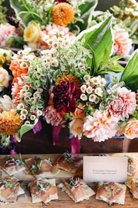 A vibrant arrangement of colorful flowers featuring dahlias, roses, and various greenery is placed above a rustic wooden table. Below the floral arrangement, neatly arranged prosciutto flatbreads topped with herbs are displayed next to a small card describing the dish.