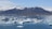 Icebergs float in a serene blue lagoon with a backdrop of snow-capped mountains under a clear sky. A small boat carrying people is navigating through the icy waters.