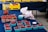A market stall displays an assortment of fresh berries. Several blue cartons filled with red raspberries are spread out on a dark blue tablecloth. A larger container holds strawberries, and there are white plastic bags containing additional berries. In the background, yellow boxes labeled 'Strawberries' are stacked, adding a bright contrast to the scene.