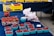 A market stall displays an assortment of fresh berries. Several blue cartons filled with red raspberries are spread out on a dark blue tablecloth. A larger container holds strawberries, and there are white plastic bags containing additional berries. In the background, yellow boxes labeled 'Strawberries' are stacked, adding a bright contrast to the scene.