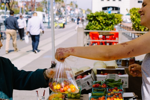 A person handing a plastic bag filled with colorful fresh tomatoes to another at a market stall. Various fresh produce, including bell peppers and more tomatoes, are displayed in containers on the stall. People can be seen walking in the background, and bright outdoor seating is visible nearby.