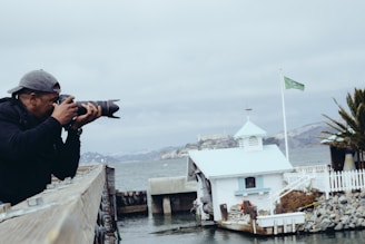 A person wearing a cap holds a professional camera, focusing intently on capturing a photograph. They stand on a wooden dock or bridge. In the background, a small white and blue building with a green flag is near the water, with distant hills and cloudy sky visible.
