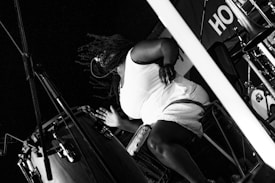 A musician is energetically playing a drum, captured in black and white. The scene includes various musical instruments and equipment, with the focus on the person immersed in the rhythm. The image conveys a sense of motion, with the person's attire and hair adding to the dynamic feel.