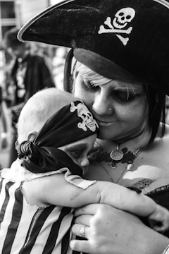 A person dressed in pirate attire is holding a baby, also dressed with a pirate theme, complete with a bandana featuring a skull and crossbones. The scene conveys a sense of affection and care.