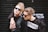 Two individuals with fashionable, edgy clothing and hairstyles, both wearing sunglasses. One is wearing a black outfit with a faux fur jacket and the other wears a black and white shirt with the word 'WHATEVER' printed on it. The background is a dark shutter with some blurred text.