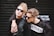 Two individuals with fashionable, edgy clothing and hairstyles, both wearing sunglasses. One is wearing a black outfit with a faux fur jacket and the other wears a black and white shirt with the word 'WHATEVER' printed on it. The background is a dark shutter with some blurred text.