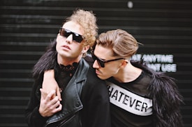 Two individuals with fashionable, edgy clothing and hairstyles, both wearing sunglasses. One is wearing a black outfit with a faux fur jacket and the other wears a black and white shirt with the word 'WHATEVER' printed on it. The background is a dark shutter with some blurred text.