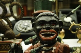 A vintage metal bank featuring the figure of a caricatured person with exaggerated facial features and holding a bowl. Behind this figure, there is an old rotary dial telephone and various other antique items including a toy dog. The surroundings are cluttered with objects, creating a sense of nostalgia.