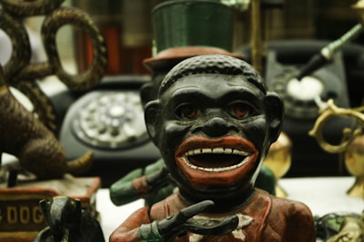 A vintage metal bank featuring the figure of a caricatured person with exaggerated facial features and holding a bowl. Behind this figure, there is an old rotary dial telephone and various other antique items including a toy dog. The surroundings are cluttered with objects, creating a sense of nostalgia.