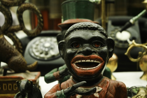 A vintage metal bank featuring the figure of a caricatured person with exaggerated facial features and holding a bowl. Behind this figure, there is an old rotary dial telephone and various other antique items including a toy dog. The surroundings are cluttered with objects, creating a sense of nostalgia.