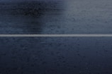 A wet road surface with a single white line running horizontally across the middle. Rain droplets and puddles are visible on the dark asphalt, creating a glossy texture.