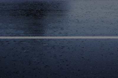 A wet road surface with a single white line running horizontally across the middle. Rain droplets and puddles are visible on the dark asphalt, creating a glossy texture.