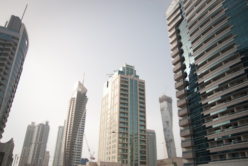 A cityscape featuring modern skyscrapers with sleek architectural designs under a clear sky. The buildings vary in shape and size, showcasing contemporary glass facades and geometric patterns. A sense of urban development is highlighted with cranes visible in the background, indicative of ongoing construction.