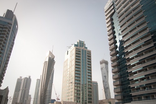A cityscape featuring modern skyscrapers with sleek architectural designs under a clear sky. The buildings vary in shape and size, showcasing contemporary glass facades and geometric patterns. A sense of urban development is highlighted with cranes visible in the background, indicative of ongoing construction.