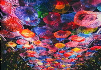 An overhead view of a colorful glass ceiling installation, featuring a variety of vibrant, translucent floral shapes. The artwork is composed of numerous glass pieces in different forms and sizes, creating a kaleidoscope effect with hues of red, blue, yellow, green, and purple.