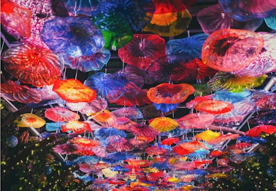 An overhead view of a colorful glass ceiling installation, featuring a variety of vibrant, translucent floral shapes. The artwork is composed of numerous glass pieces in different forms and sizes, creating a kaleidoscope effect with hues of red, blue, yellow, green, and purple.