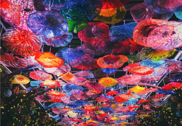 An overhead view of a colorful glass ceiling installation, featuring a variety of vibrant, translucent floral shapes. The artwork is composed of numerous glass pieces in different forms and sizes, creating a kaleidoscope effect with hues of red, blue, yellow, green, and purple.