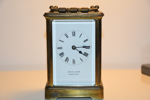 An antique brass carriage clock with a rectangular shape and a white clock face displaying Roman numerals. The casing has a handle on top, and the name 'REED & SON CAMBRIDGE' is visible on the face.