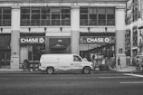 A street scene featuring a large building with prominent Chase bank signage. In front of the building, a white van with 'FLOWERS, INC.' branding is parked on the road. Pedestrians are visible walking along the sidewalk, and a few are gathered near the bank entrance. The image is rendered in black and white, giving it a vintage feel.
