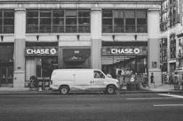 A street scene featuring a large building with prominent Chase bank signage. In front of the building, a white van with 'FLOWERS, INC.' branding is parked on the road. Pedestrians are visible walking along the sidewalk, and a few are gathered near the bank entrance. The image is rendered in black and white, giving it a vintage feel.