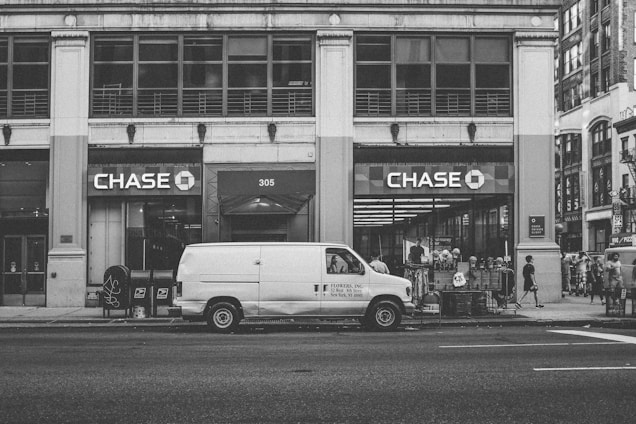 A street scene featuring a large building with prominent Chase bank signage. In front of the building, a white van with 'FLOWERS, INC.' branding is parked on the road. Pedestrians are visible walking along the sidewalk, and a few are gathered near the bank entrance. The image is rendered in black and white, giving it a vintage feel.