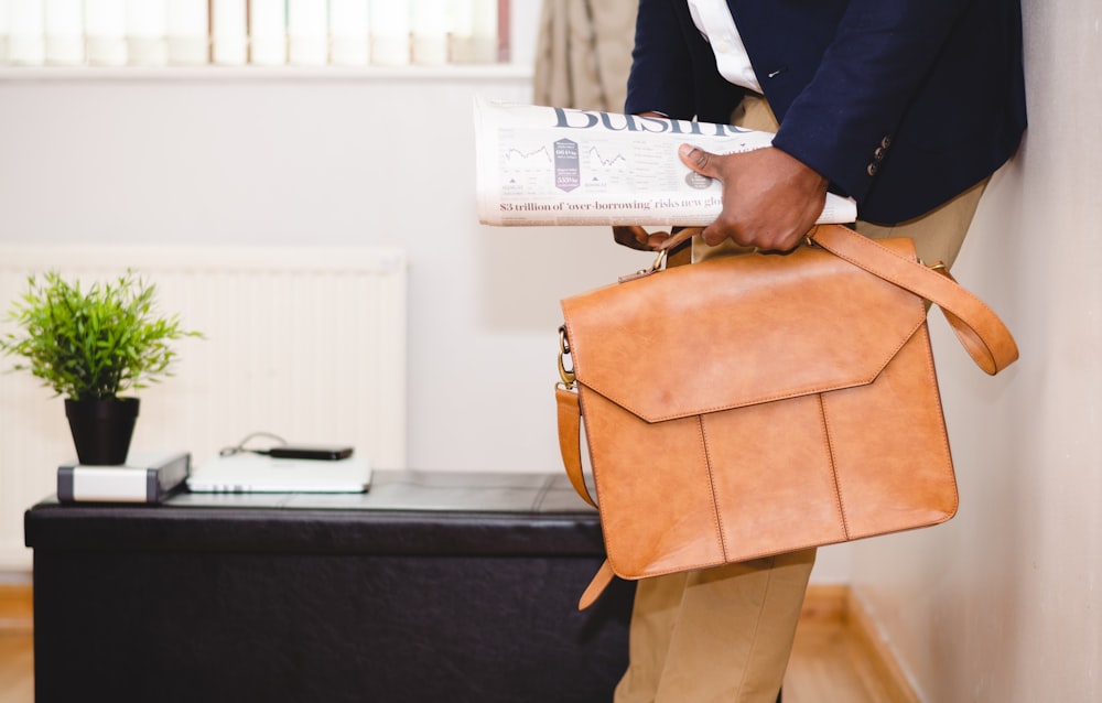 homme tenant un sac en cuir marron appuyé sur un mur blanc