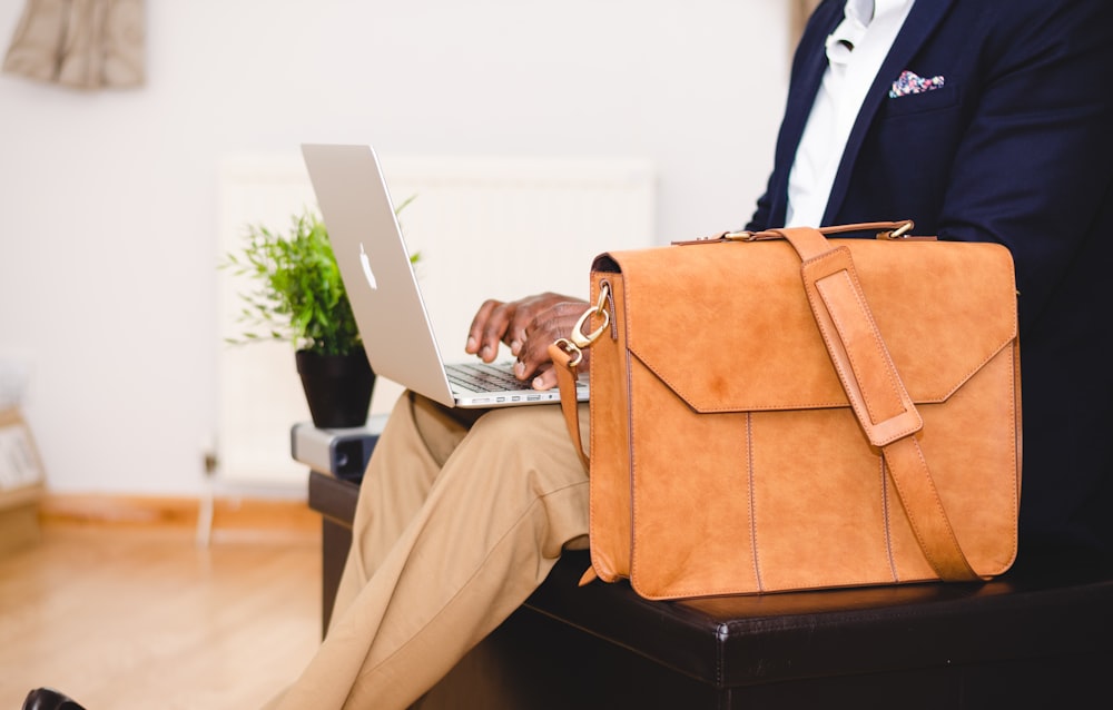 person using MacBook Pro beside brown leather briefcase