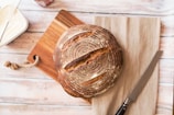 A loaf of round, artisan-style bread with a crusty surface is placed on a wooden cutting board with a rope handle. A serrated knife with a black handle lies next to the bread on a beige cloth. To the left, there is a container with a lid partially visible, most likely containing butter or spread, with a knife resting on it. The background consists of a light-colored wooden surface.