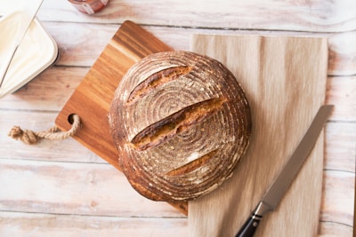 A loaf of round, artisan-style bread with a crusty surface is placed on a wooden cutting board with a rope handle. A serrated knife with a black handle lies next to the bread on a beige cloth. To the left, there is a container with a lid partially visible, most likely containing butter or spread, with a knife resting on it. The background consists of a light-colored wooden surface.