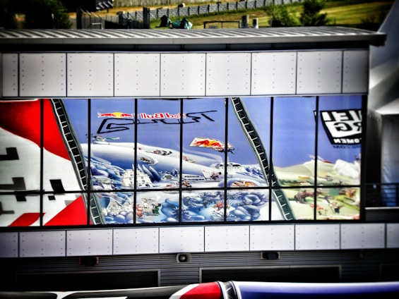 A reflective building facade displays a vibrant mural featuring dynamic and colorful elements such as clouds, people, aircraft, and various scenes. The reflection is seen in large rectangular window panes, offering a fragmented yet intriguing visual effect. The artwork includes text and logos, enhancing the energetic and playful representation.