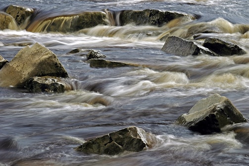 Water flows rapidly over and around large, jagged rocks in a stream or river. The movement creates soft, blurred lines in the water, contrasting with the sharp, defined edges of the stones.