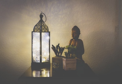 An intricately designed lantern emits a soft glow, illuminating a small statue of Buddha and two potted plants. The setup is placed on a wooden surface against a plain, light-colored wall, creating a peaceful and serene atmosphere.