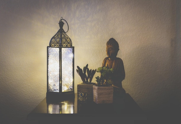 An intricately designed lantern emits a soft glow, illuminating a small statue of Buddha and two potted plants. The setup is placed on a wooden surface against a plain, light-colored wall, creating a peaceful and serene atmosphere.