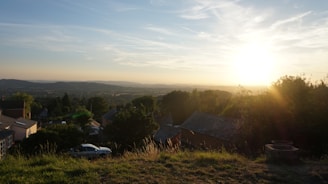 A scenic view at sunset over a small settlement in a hilly area. The sun is low on the horizon, casting a warm golden light over the landscape. There are a few houses and buildings surrounded by trees and greenery. The sky is clear with some scattered clouds, and the overall scene exudes tranquility and beauty.