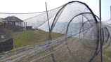 A large fishing net structure is set up outdoors on a grassy area near the water. The net is composed of multiple circular frames, creating a tunnel-like appearance. In the background, there are wooden pavilions and the sea, with a clear sky overhead.