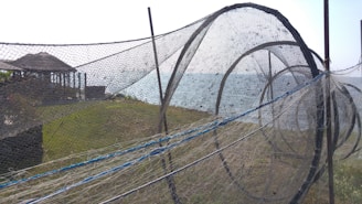 A large fishing net structure is set up outdoors on a grassy area near the water. The net is composed of multiple circular frames, creating a tunnel-like appearance. In the background, there are wooden pavilions and the sea, with a clear sky overhead.