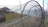 A large fishing net structure is set up outdoors on a grassy area near the water. The net is composed of multiple circular frames, creating a tunnel-like appearance. In the background, there are wooden pavilions and the sea, with a clear sky overhead.