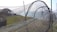A large fishing net structure is set up outdoors on a grassy area near the water. The net is composed of multiple circular frames, creating a tunnel-like appearance. In the background, there are wooden pavilions and the sea, with a clear sky overhead.