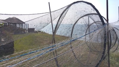 A large fishing net structure is set up outdoors on a grassy area near the water. The net is composed of multiple circular frames, creating a tunnel-like appearance. In the background, there are wooden pavilions and the sea, with a clear sky overhead.