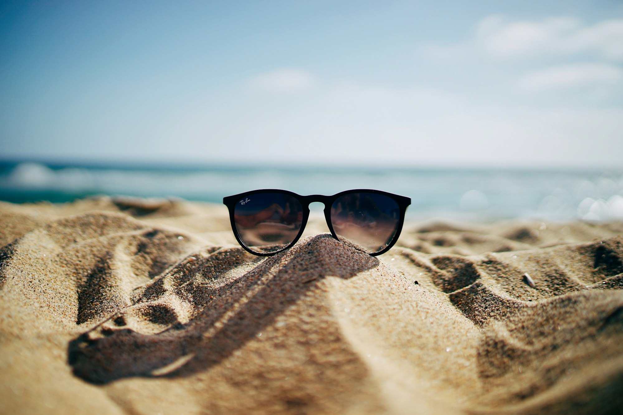 Paire de lunettes de soleil Ray Ban noires posées sur le sable devant la mer
