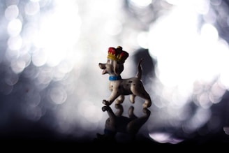 A small figurine of a spotted dog wearing a red and gold crown is positioned against a backdrop of blurred lights, creating a bokeh effect. The dog appears joyful with its mouth open, and it is standing on a reflective surface that mirrors its image.
