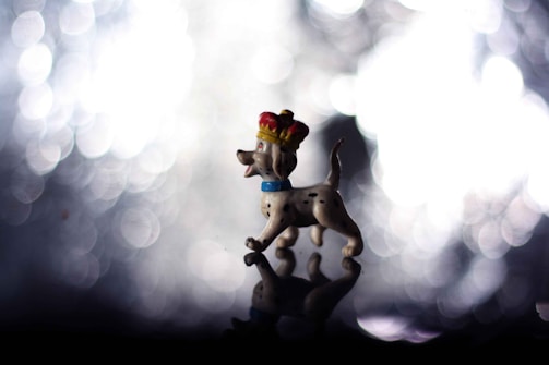 A small figurine of a spotted dog wearing a red and gold crown is positioned against a backdrop of blurred lights, creating a bokeh effect. The dog appears joyful with its mouth open, and it is standing on a reflective surface that mirrors its image.