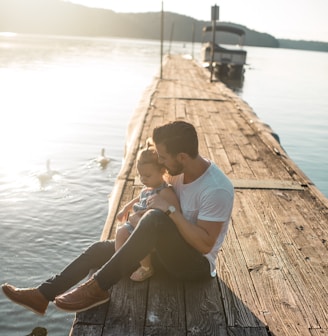 man and girl sitting on brown dock near boat and two white ducks during daytime