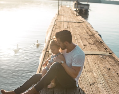 man and girl sitting on brown dock near boat and two white ducks during daytime