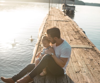 man and girl sitting on brown dock near boat and two white ducks during daytime