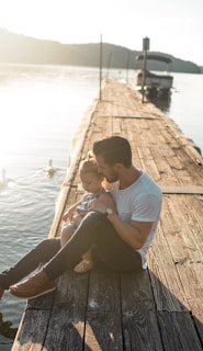 man and girl sitting on brown dock near boat and two white ducks during daytime